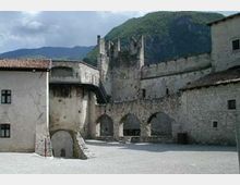 Cortile interno di una fortificazione in pietra con alte mura merlate e una torre centrale, con serie di archi lungo il perimetro. Sullo sfondo si vedono edifici con tetti in tegole e montagne boscose.