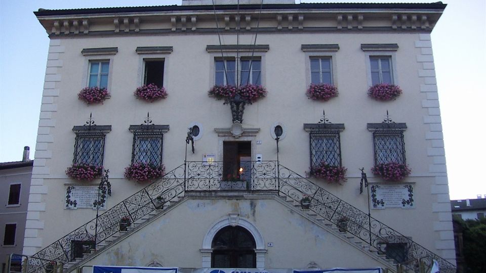 Facciata di un edificio civico storico a tre piani con finestre simmetriche decorate da fioriere, ingresso centrale sopra una scalinata doppia con ringhiere in ferro battuto e piccolo campanile con campana sul tetto. Nella parte bassa sono visibili striscioni con scritte e un portale ad arco.
