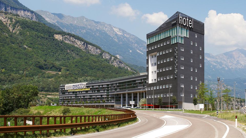 Edificio alberghiero moderno con torre alta scura e insegna “hotel”, affiancato da un lungo corpo basso con molte finestre. La struttura si trova lungo una strada curva in un’area verde ai piedi di montagne rocciose e boscose.