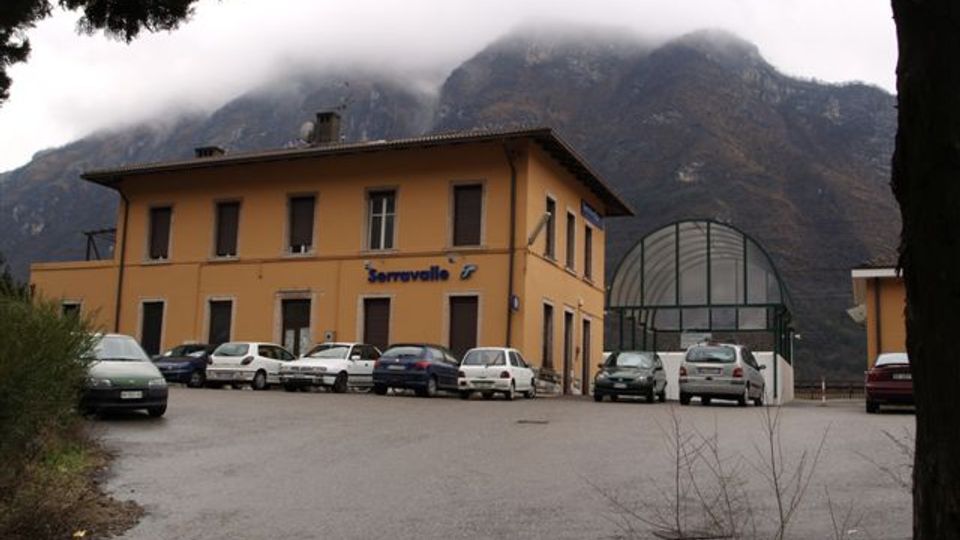 Edificio basso in stile stazione ferroviaria, color ocra, con la scritta “Serravalle” sulla facciata e un ampio piazzale asfaltato con auto parcheggiate davanti. Sullo sfondo si vedono montagne ripide parzialmente coperte da nuvole e, a destra, una struttura a serra con tetto arcuato in vetro.