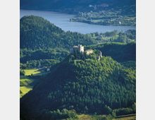 Castello in pietra con torre cilindrica su una collina boscosa, circondato da fitte foreste e campi coltivati. Sullo sfondo si vede un grande lago con piccoli centri abitati lungo la riva e pendii montuosi.
