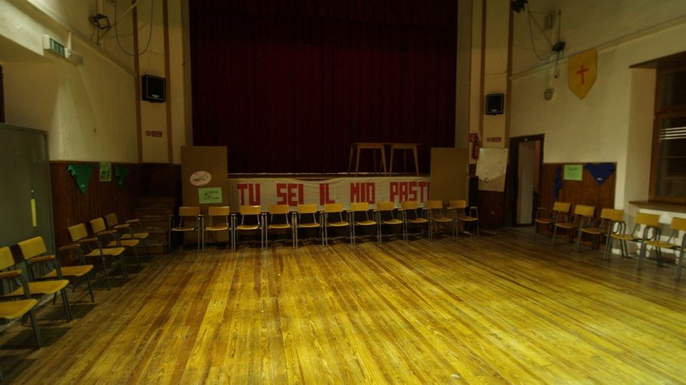 Sala polivalente con ampio pavimento in legno e sedie disposte lungo le pareti, di fronte a un palco rialzato con grande tenda scura. Sulle pareti laterali sono presenti luci, altoparlanti e decorazioni, con una scritta sul fronte del palco.