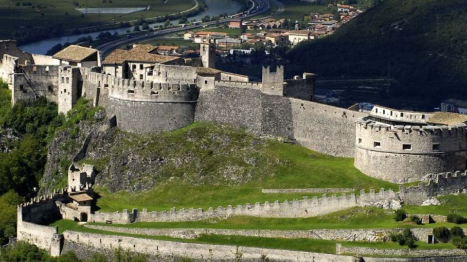 Veduta aerea di un grande castello medievale in pietra con bastioni, torri e mura merlate, disposto su più livelli lungo una collina boscosa. In primo piano si vedono le fortificazioni esterne e un prato interno, con la valle e un centro abitato sullo sfondo.