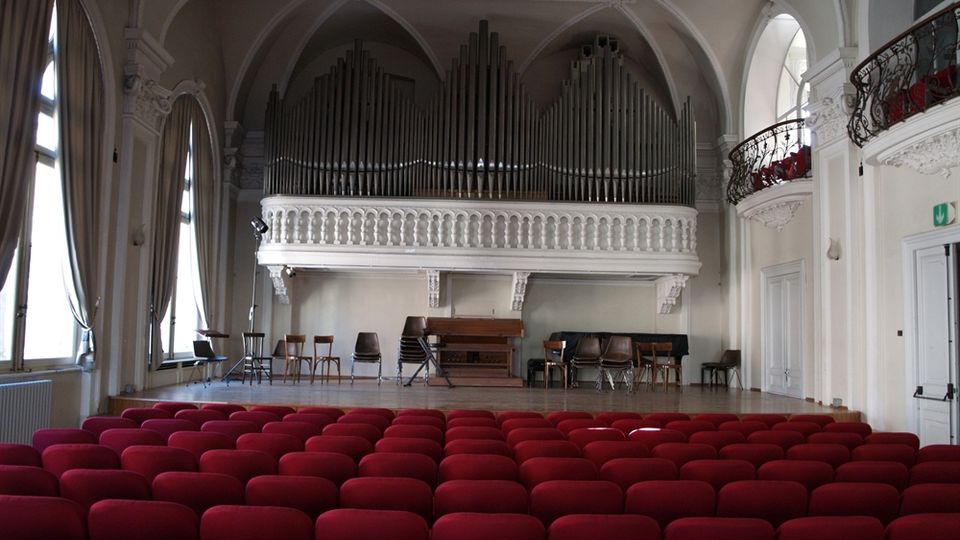 Interno di una sala da concerto con file di poltrone rosse e palco in legno, sormontato da una grande cantoria con organo a canne. L’ambiente presenta soffitto a volte, grandi finestre con tende e una balconata laterale con ringhiera decorata.