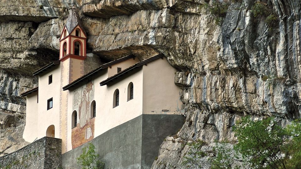 Piccola chiesa con campanile e facciata affrescata incastonata sotto una grande parete di roccia stratificata. L’edificio chiaro ha tetti spioventi e finestre ad arco, con vegetazione alla base della scogliera.