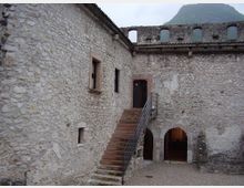 Cortile interno di una struttura storica in pietra con scala esterna in mattoni e corrimano metallico che conduce a una porta sopraelevata. Al piano terra si vedono due archi d’ingresso e, sullo sfondo, un muro alto con aperture ad arco e una montagna oltre la sommità.