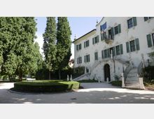 Facciata di una villa storica intonacata di bianco con persiane verdi, ingresso ad arco e doppia scalinata esterna con balaustre e statue. Davanti si vede un cortile con ghiaia, siepi potate e alti cipressi in un giardino alberato.