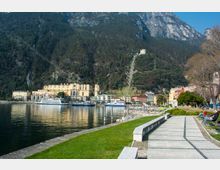 Lungolago con passeggiata lastricata e prato lungo la riva, con pontili e battelli attraccati davanti a edifici sul waterfront. Sullo sfondo si alzano montagne boscose e rocciose attraversate da una funicolare che sale verso una struttura in cima al pendio.