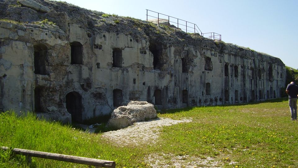 Lungo edificio in cemento e pietra, parzialmente scavato nella roccia, con numerose aperture ad arco e rettangolari simili a feritoie lungo la facciata. Davanti si vede un prato con un sentiero ghiaioso e, in alto, una ringhiera metallica sul bordo della struttura sotto un cielo sereno.