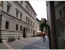 Strada pedonale stretta tra alti edifici in pietra, con facciata bugnata e finestre ad arco su un grande palazzo storico. Pavimentazione in lastre, panchine e biciclette lungo il muro, con alberi e ombrelloni di un locale visibili sullo sfondo.