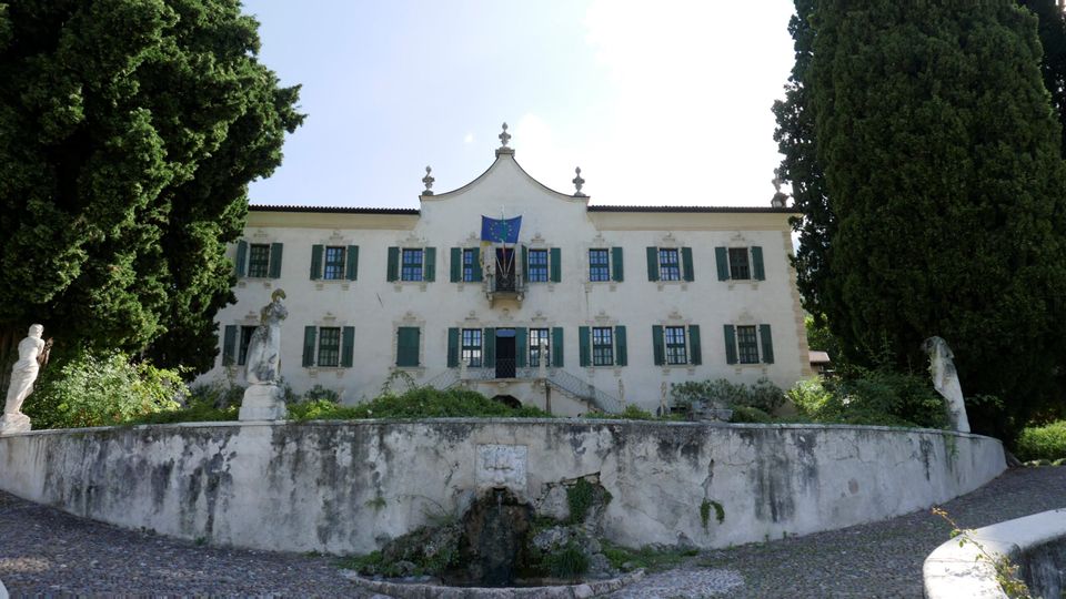 Facciata di una villa storica intonacata di bianco con persiane verdi e frontone centrale, affacciata su un giardino rialzato con muretto e piccola fontana. Due grandi alberi ai lati incorniciano l’edificio, con statue in pietra lungo il bordo del giardino.