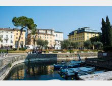 Veduta di un piccolo porto urbano con barche ormeggiate in una darsena circolare in muratura. Sullo sfondo si vedono edifici cittadini a più piani dai colori chiari, un’area verde con alberi e un ampio cielo sereno.