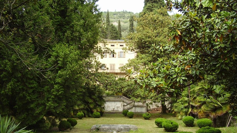 Giardino di una villa con prato e siepi potate, circondato da alberi sempreverdi e magnolie. Sullo sfondo si vede un edificio chiaro a più piani e una scalinata con balaustra in pietra, con colline alberate oltre la proprietà.