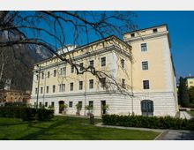 Edificio storico a più piani con facciata giallo chiaro e basamento in pietra bianca, finestre regolari e un grande portone ad arco. Davanti si estende un prato curato con siepi e vasi, attraversato da vialetti, con rami di un albero in primo piano e colline rocciose sullo sfondo.