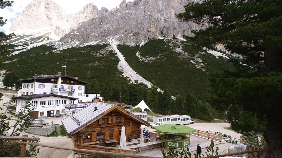 Rifugio e piccole strutture in legno e muratura con terrazza esterna in una radura di montagna, circondati da boschi di conifere. Sullo sfondo si alzano alte pareti rocciose e cime frastagliate con chiazze di neve.
