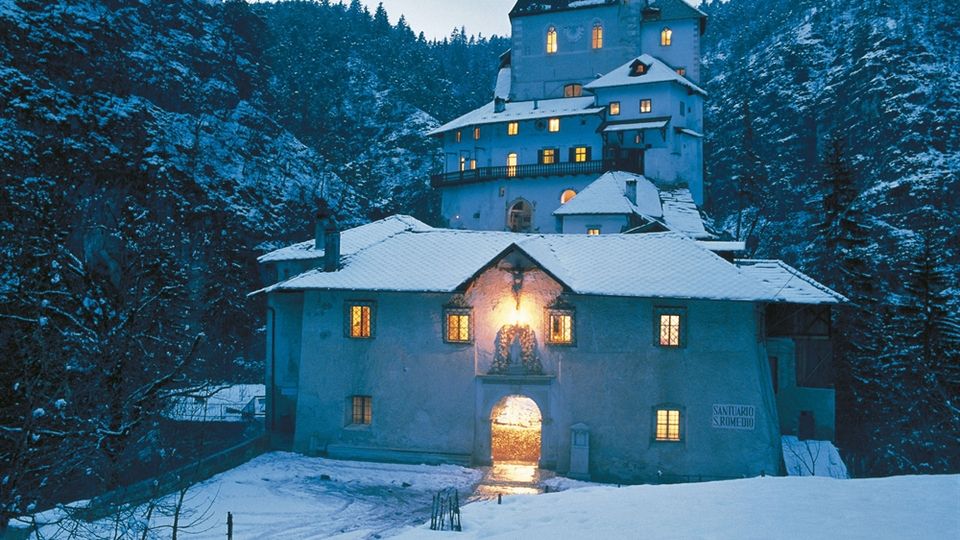 Santuario e complesso monastico in pietra su un pendio boscoso, con tetti innevati e finestre illuminate al crepuscolo. L’edificio principale presenta una torre con orologio e campanile, circondato da montagne e alberi coperti di neve.