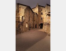 Vicolo in un borgo storico con edifici in pietra e intonaco consumato, finestre piccole e muri parzialmente crollati. In fondo si vede un arco che attraversa il passaggio, con strada in ghiaia tra le facciate antiche.