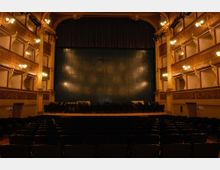 Interno di un teatro all’italiana visto dalla platea, con file di poltrone scure rivolte verso un ampio palcoscenico con sipario e fondale scuro. Ai lati si notano più ordini di palchi decorati in legno con luci a parete e dettagli ornamentali.