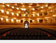 Interno di un teatro storico con platea di sedie scure e tre ordini di palchi curvilinei decorati in tonalità oro, illuminati da lampadari e appliques. Al centro si vede un grande arco con tendaggi che conduce a un ingresso sul fondo.