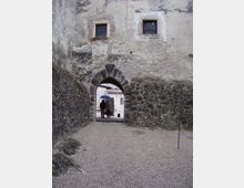 Passaggio in ghiaia che conduce a un arco in pietra all’ingresso di un edificio storico con muri in intonaco consumato e base in pietre a vista. Sopra l’arco si vedono due piccole finestre quadrate, mentre oltre l’arco appare un cortile interno con altre strutture in pietra.