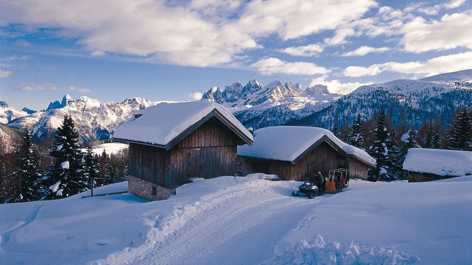 Baita e piccoli edifici in legno con tetti molto innevati su un pendio alpino, circondati da abeti e neve. Sullo sfondo si vedono alte montagne innevate sotto un cielo azzurro con nuvole.