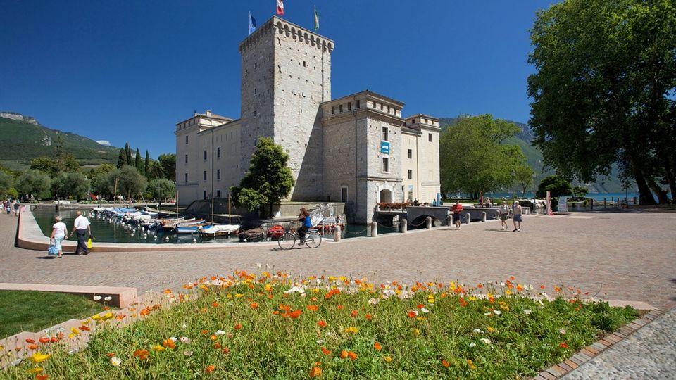 Castello in pietra con alta torre merlata affacciato su un porto con barche ormeggiate e un lungolago. In primo piano una piazza lastricata con aiuola fiorita, alberi e montagne sullo sfondo sotto cielo sereno.