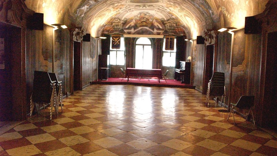 Interno di una sala storica con soffitto a volta riccamente affrescato e pareti decorate, illuminata da lampade laterali. Pavimento a scacchiera in pietra e, sul fondo, una pedana con tavolo davanti a una grande finestra ad arco.