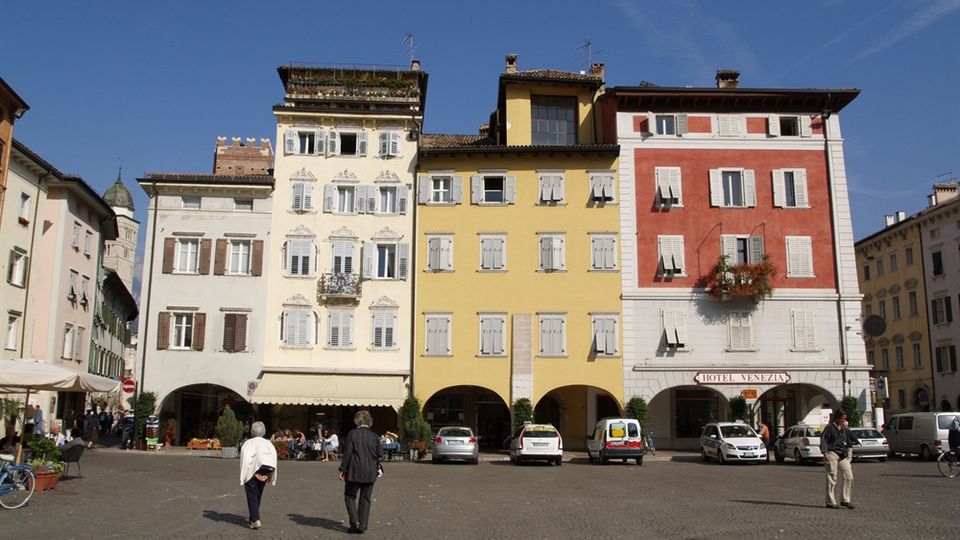 Piazza lastricata con file di edifici storici a più piani dai colori pastello (bianco, giallo e rosso), con finestre a persiane e portici ad archi al piano terra. Negozi e un’insegna “Hotel Venezia” si affacciano sulla piazza, con alcune auto parcheggiate davanti.