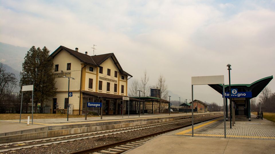 Stazione ferroviaria di Roncégno con binari in primo piano e banchine in cemento, segnaletica blu e pensiline moderne a lato. Sullo sfondo si vede l’edificio storico della stazione a due piani e un paesaggio pianeggiante con alberi sotto un cielo nuvoloso.