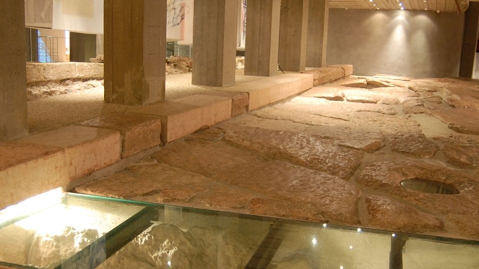 Interno di uno spazio museale sotterraneo con colonne in cemento e soffitto curvo rivestito in legno, illuminato da faretti lineari. Il pavimento presenta lastre di pietra e pannelli di vetro che permettono di vedere scavi e reperti archeologici sottostanti, con pannelli espositivi lungo il percorso.
