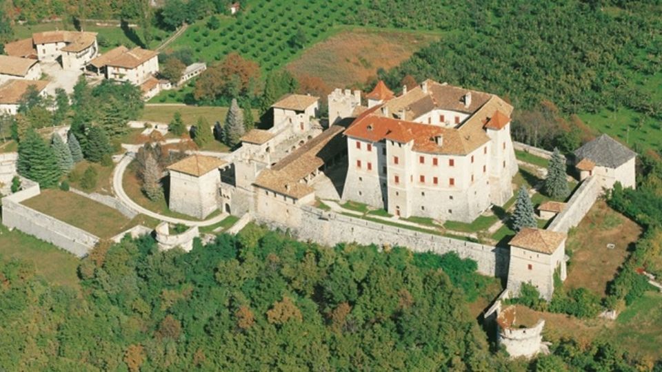 Veduta aerea di un complesso fortificato con mura in pietra e torri, con un grande edificio centrale dai tetti in tegole rosse e vari annessi. Il castello è circondato da boschi e campi coltivati su un’area collinare.