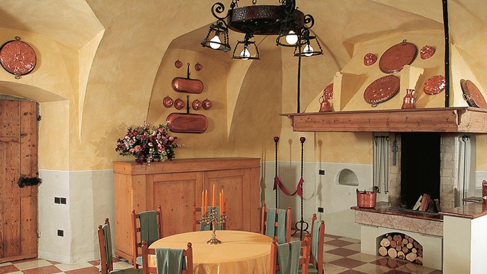 Interno rustico con soffitto a volta intonacato, pavimento a scacchi rosso e bianco e grande camino in muratura con legnaia. Al centro c’è un tavolo rotondo con sedie in legno, illuminato da un lampadario in ferro battuto, con piatti decorativi alle pareti e una credenza in legno.