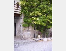 Cortile con parete in pietra di un edificio rustico, in gran parte ricoperta da una fitta pianta rampicante verde. In basso si vedono ghiaia e ciottoli, una panca in pietra con vaso di fiori e alcuni elementi decorativi, oltre a ruote di carro appoggiate al muro.