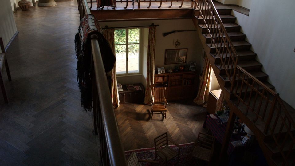 Interno di una casa a due piani visto dall’alto, con ballatoio in legno e scala con ringhiera che scende verso un soggiorno a doppia altezza. In basso si vedono pavimento in parquet, finestra alta con tende, libreria e sedute attorno a un tappeto.