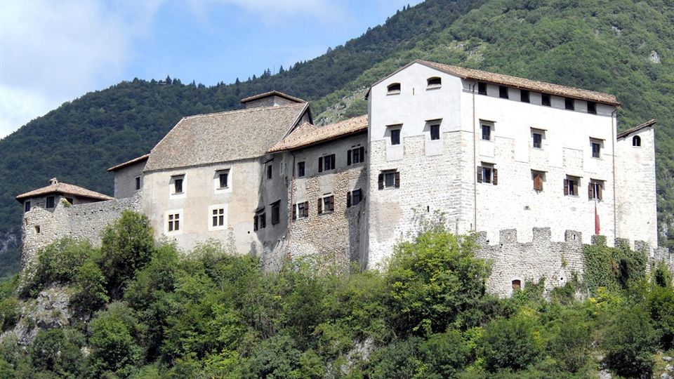 Castello in pietra con grandi mura chiare, finestre piccole e tetti in tegole, arroccato su un pendio sopra una vegetazione fitta. Sullo sfondo si vedono montagne boscose sotto un cielo parzialmente nuvoloso.