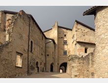 Cortile di un borgo medievale con edifici in pietra e intonaco consumato, pareti parzialmente diroccate e finestre piccole. Un passaggio ad arco collega le costruzioni, con tetti in coppi e un vialetto in pendenza tra i muri di pietra.