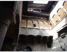 Cortile interno di un edificio storico in pietra, visto dal basso, con loggiati ad archi su colonne e ballatoi sovrapposti. In alto si nota una copertura parziale con apertura che lascia entrare luce, e sulle pareti sono presenti finestre ad arco e una lanterna sospesa.