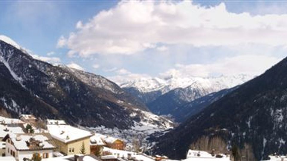 Veduta panoramica di una valle alpina innevata con montagne boscose sui lati e cime più alte sullo sfondo. In primo piano si vede un piccolo paese con case e tetti coperti di neve sotto un cielo parzialmente nuvoloso.