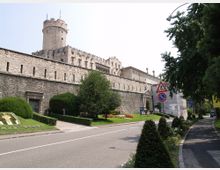 Castello in pietra con alte mura merlate e torre cilindrica, affacciato su una strada asfaltata con aiuole curate e siepi. Lungo il margine stradale si vedono segnali di attraversamento pedonale e divieto di sosta, con alberi che incorniciano la scena.