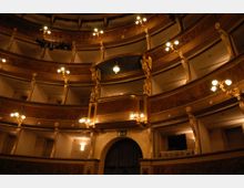 Interno di un teatro storico con più ordini di palchi curvi sovrapposti, ricchi di decorazioni dorate e statue, illuminati da lampadari e applique. In basso si vede un grande arco con tendaggio scuro che conduce verso il palco o un corridoio.