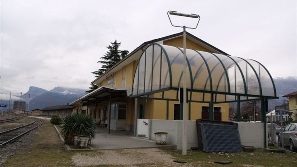 Piccola stazione ferroviaria con edificio giallo e marciapiede affiancato ai binari, con una pensilina ad arco in struttura metallica e pannelli traslucidi sul lato. Intorno si vedono un piazzale sterrato, alcune piante e montagne sullo sfondo sotto un cielo nuvoloso.