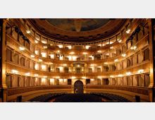 Interno di un teatro all’italiana con platea e più ordini di palchi sovrapposti disposti in forma semicircolare. Le balconate sono ornate con decorazioni dorate e illuminate da numerose lampade lungo i parapetti, sotto un soffitto affrescato.