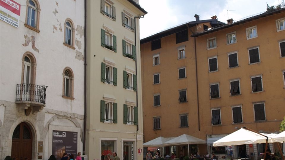 Piazza cittadina lastricata con edifici storici dai toni chiari e ocra, con finestre a persiane e una torre in pietra merlata con bandiera. Sul lato della piazza ci sono ombrelloni e tavolini all’aperto, vasi di piante e piccoli banchi.
