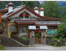 Casa alpina decorata con intagli in legno, motivi dipinti e balconcino con fiori, con tetto a scandole e camini con cappelli rossi. Davanti ci sono una staccionata, un ingresso con due pilastri scolpiti a forma di volti e un piccolo giardino, sullo sfondo boschi e montagne.