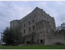 Grande castello in pietra su un’altura, con mura massicce, finestre strette e una torre cilindrica laterale. In primo piano si vedono un bastione con tetto a padiglione, un arco d’ingresso, prato e alberi sotto un cielo nuvoloso.