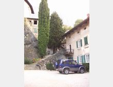 Cortile di un piccolo borgo montano con case in pietra e intonaco, finestre con scuri verdi e un balcone in ferro. Un muretto in pietra e una scalinata salgono tra alti cipressi e una grande roccia, con auto parcheggiata in primo piano.