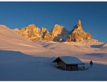Paesaggio alpino invernale con ampie distese di neve e una piccola baita in legno semisepolta in primo piano. Sullo sfondo si stagliano cime rocciose illuminate dalla luce calda del sole contro un cielo blu limpido.