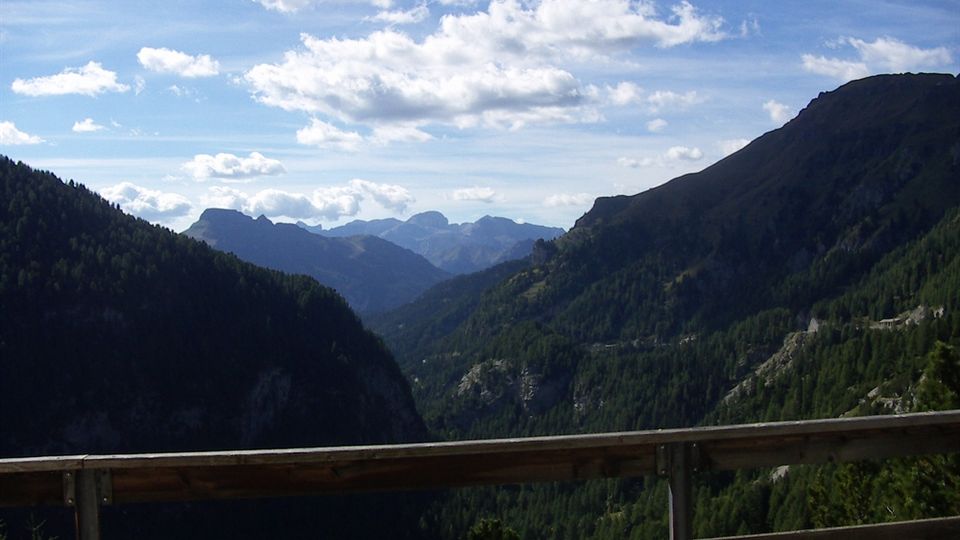 Veduta panoramica di una valle montana con pendii boscosi e cime rocciose in lontananza sotto un cielo azzurro con nuvole sparse. In primo piano si vede una staccionata in legno, come da belvedere o strada panoramica.