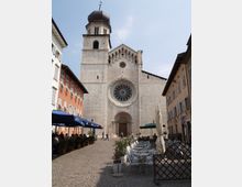 Facciata di una chiesa in pietra chiara con grande rosone centrale, portale ad arco e campanile laterale con cupola scura. Davanti si apre una piazza lastricata fiancheggiata da edifici e tavolini di bar con ombrelloni.