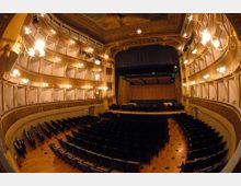 Interno di un teatro all’italiana con platea di poltrone scure e palchi sovrapposti decorati e illuminati da lampade a parete. In fondo si vede il palco con sipario e struttura scenica sotto un soffitto affrescato.